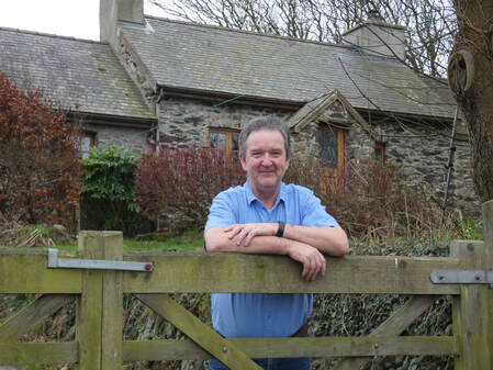 Bruce Clifton resting on his arms, leaning against a gate, standing in front of a stone cottage in Wales on a winters day.